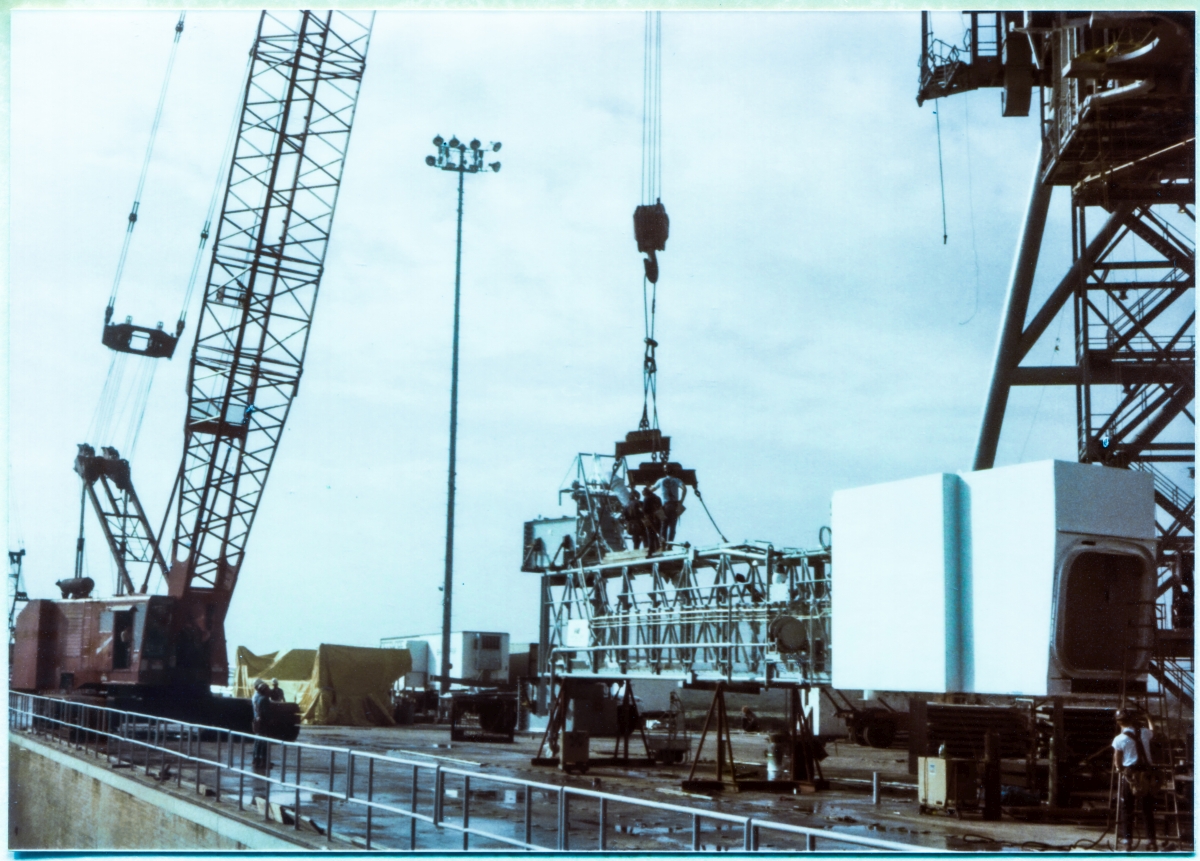 Image 130. OAA Lift 2 of 9. At Space Shuttle Launch Complex 39-B, Kennedy Space Center, Florida, the Orbiter Access Arm (OAA) can be seen sitting on its support stanchions, on top of the Pad Deck, while Union Ironworkers from Local 808 working for Ivey Steel Erectors attach lifting gear to it. The crane which will lift the OAA to its final working position at Elevation 200'-0” on Side 1 of the Fixed Service Structure in a few more minutes is visible very near the west wall of the Flame Trench, in the lower left corner of the photograph. A close look at the run of handrail which guards the Flame Trench, along with the aspect of the Flame Trench Wall itself, as well as the perceived elevation of the OAA's Bottom Truss Chord, reveals that the photograph was taken from a position standing among the SSW Spray Nozzles along the Flame Deflector Crest, not quite midway along its length, a bit closer to the west side of the Trench than its east side. This is a very unusual viewpoint for any image ever taken of the Pad, and was a bit hazardous to access owing to the difficulty of traversing the line of Spray Nozzles without getting tripped-up doing so, in conjunction with the consequences of a fall down the Fondu Fyre covered Flame Deflector Slope on either side of its Crest. Photo by James MacLaren.